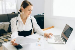 Une femme à son bureau examine des rapports commerciaux sur un ordinateur portable, tenant des papiers dans un environnement de bureau moderne.